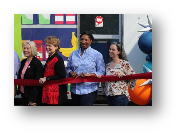 Ribbon Cutting of the UALR University District Mobile Market - March 2026. Four women, smiling, holding a wide red ribbon prior to a ribbon cutting ceremony. The mobile market is in the background with images of fruit on the exterior. There is a blue, turquoise, orange, and silver balloon sculpture next to the market door.
